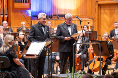 dwóch mężczyzn w eleganckich smokingach trzyma w ręku notatki i przemawia w stronę publiczności, stojąc na środku estrady, za nimi siedzi orkiestra