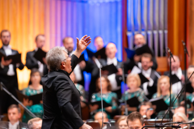 Zdjęcie dyrygenta z ręką w górze spoglądającego w górę, w stronę stojącego przed nim chóru