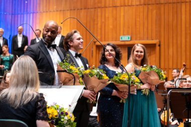 dwie kobiety i dwoje mężczyzn w eleganckich, wieczorowych strojach z bukietami kwiatów w rękach stoją na środku estrady przed orkiestrą, zwróceni w stronę publiczności