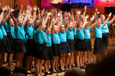 chór dziecięcy w niebieskich koszulkach, śpiewający z uniesionymi ku górze rękami
