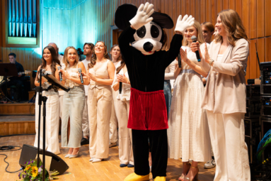 chór studnetów w jasnych strojach, przed którymi stoi maskotka Myszka Miki machająca w stronę publiczności