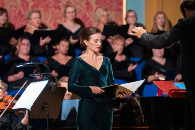 kobieta w zielonej, welurowej sukience spoglądająca w teczkę z nutami, za nia chór i orkiestra