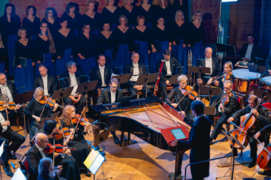 orkiestra i chór na estradzie widziani z góry. Na środku, pomiędzy nimi stoi fortepian